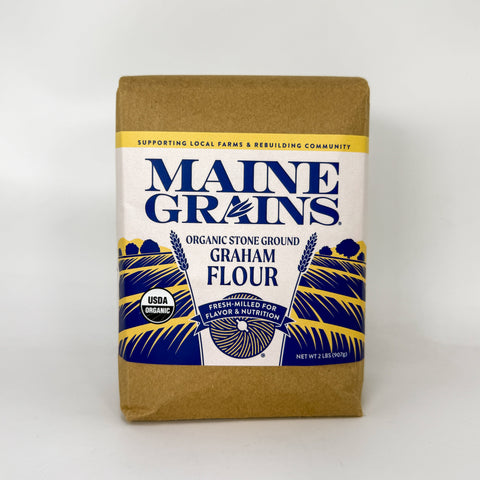 A bag of Maine Grains Organic Stone Ground Graham Flour, packaged in a brown kraft paper bag with a white, blue, and yellow label.