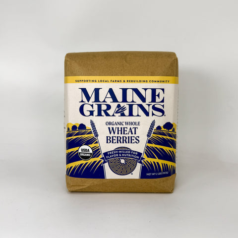 A package of Maine Grains Organic Whole Wheat Berries on a clean white background. The packaging features a brown kraft paper top and bottom with a white center label containing blue and yellow graphics.