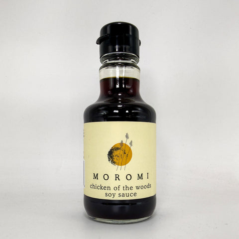 A bottle of Moromi chicken of the woods soy sauce with a cream label and black cap, photographed on a light gray background.
